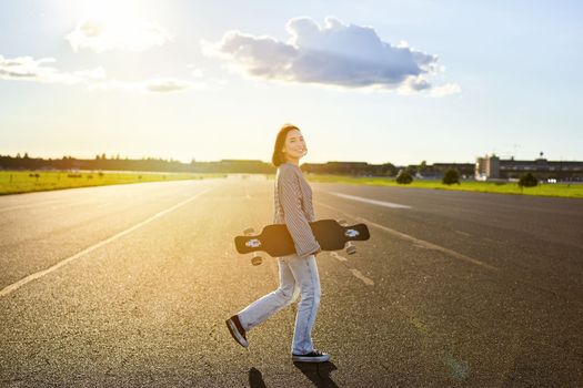 Asian girl with skateboard standing on road during sunset. Skater posing with her long board, cruiser deck during training.