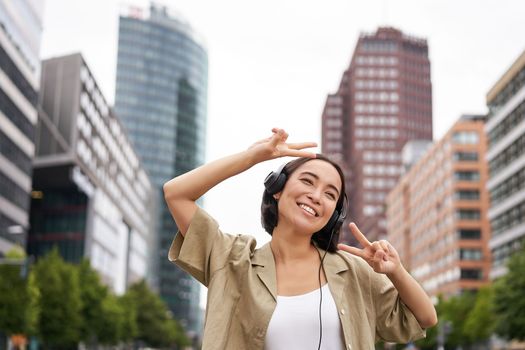 Happy asian woman in headphones, listening music and dancing on street of city centre, smiling with hands up.