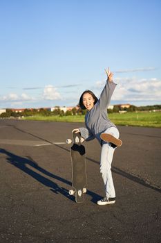 Vertical shot of happy asian skater girl, jumping, standing with skateboard and smiling. Woman skating on longboard and having fun.