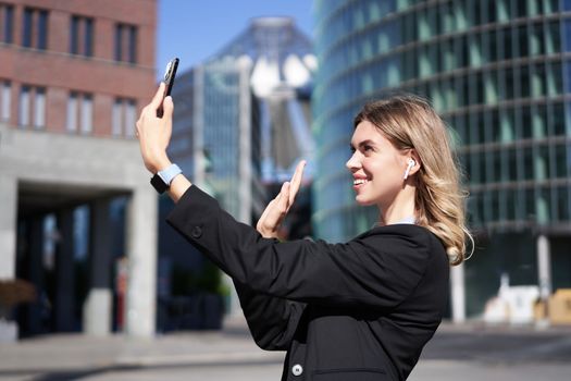 Portrait of businesswoman wave her hand at mobile phone camera, waves hand during video chat, stands in suit in city center outdoors.