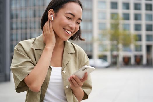 Smiling asian girl listens music in wireless headphones, looks at her phone, choosing music or podcast. Young woman calling someone, using headphones.