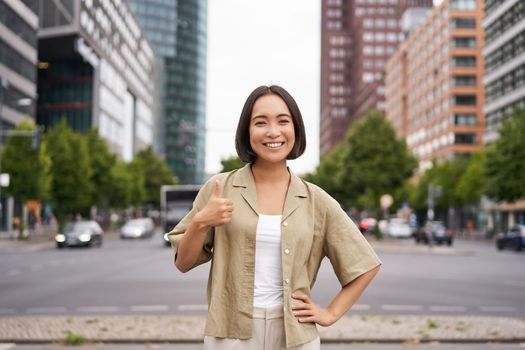 Enthusiastic city girl, shows thumbs up in approval, looking upbeat, say yes, approves and agrees, stands on street.