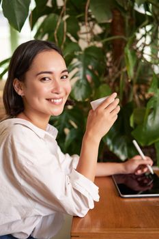 Vertical shot of young asian woman with digital tablet and graphic pen, drawing, scratching while sitting in cafe with cup of coffee.