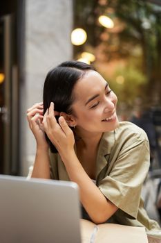 Vertical shot of beautiful asian girl sits with laptop in cafe, tucks hair strand behind ear and smiles.