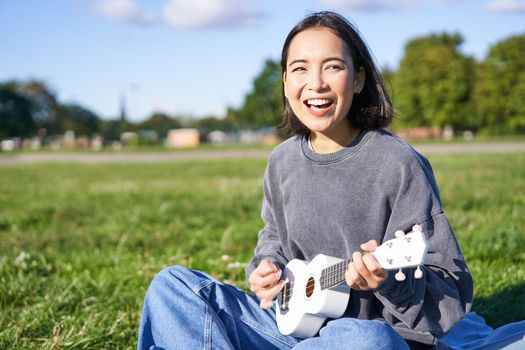 Portrait of asian girl student, playing ukulele and singing in park, sitting alone on blanket and enjoying making music.