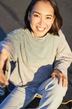 Selfie of beautiful asian girl smiling, taking photo on smartphone while sitting on skateboard outdoors.
