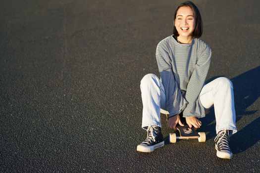 Freedom and happiness. Cute smiling asian girl, sits on skateboard on sunny spring day. Happy laughing skater enjoying cruising on longboard.