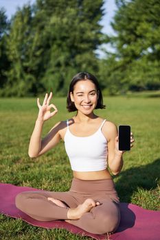 Vertical shot of asian woman shows okay sign and smartphone screen, recommending yoga training online, meditation app, doing exercises on fresh air in park.