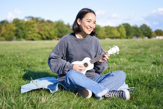 Beautiful asian girl sitting in park, playing ukulele and singing, relaxing outdoors on sunny spring day.