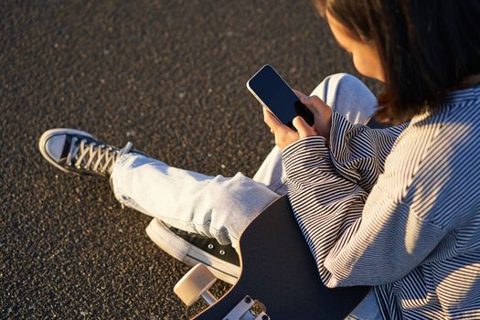 Close up of skater teen girl sits on skateboard, types message on smartphone, looks at mobile phone screen.