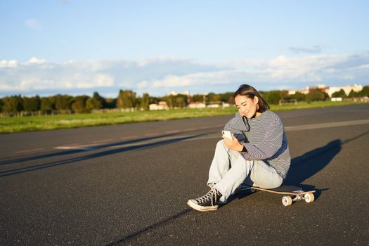 Portrait of young korean girl sitting on her skateboard on road, looking at smartphone, chatting on mobile app.