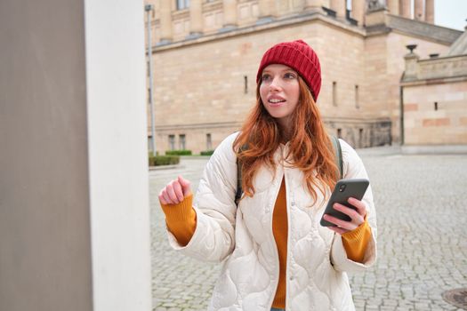 Tourism and sightseeing concept. Young redhead woman, tourist walks around city, looks at her smartphone app and at history stand, explores adventures.