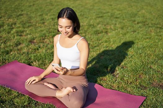 Asian girl follows yoga training app, looking at smartphone, meditating and doing exercises on fresh air in park.