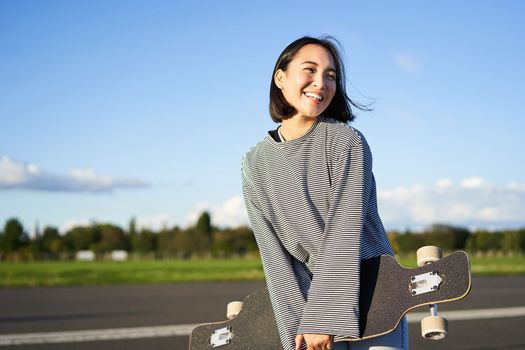 Portrait of asian woman with longboard. Korean girl skating, holding skateboard in hands, posing on road, smiling and looking aside.