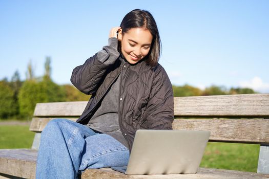 Young brunette girl sits in park with laptop, watches video or browses internet, rests outdoors on bench in sunny park.