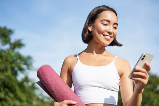 Portrait of smiling asian woman with yoga mat, looking at her smartphone and reading on application, standing in park wearing sport uniform.