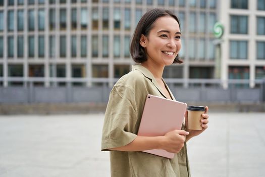 Portrait of happy asian girl with tablet and coffee, walking on street with pleased smile.