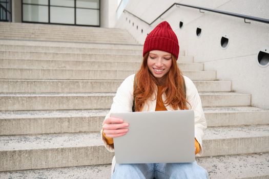 Smiling redhead girl, student sits on stairs outdoors and uses laptop, connects to public wifi in city and works on project, uses internet on computer.