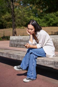 Portrait of asian girl sitting with smartphone feeling sad, looking gloomy and frustrated, waiting for a call outdoors in park.