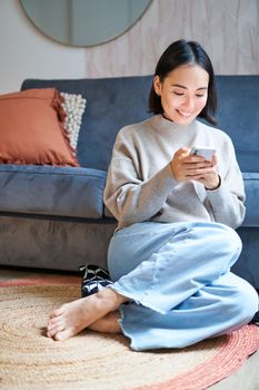 People and technology. Smiling asian woman sitting at home, using her mobile phone, typing message, browing internet or shopping online from smartphone app.