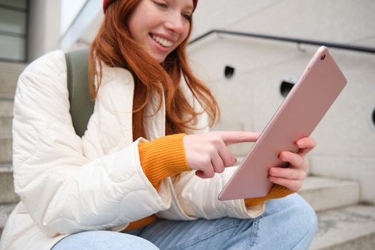 Happy stylish redhead girl, student in red hat, holds digital tablet, uses social media app, searches something online, connects to wifi.