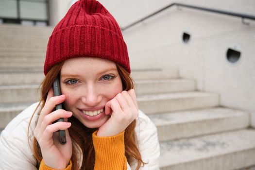 Young stylish redhead girl in red hat, sits on street and talks on mobile phone, has telephone conversation, rings her friend while relaxes outdoors.