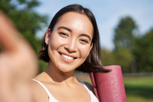 Happy asian fitness girl takes selfie with rubber yoga mat in park. Healthy young sportswoman makes photo of herself during workout.