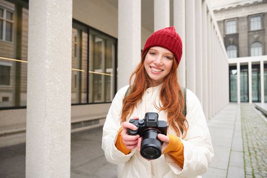 Smiling redhead girl photographer, taking pictures in city, makes photos outdoors on professional camera. Young talent and hobby concept