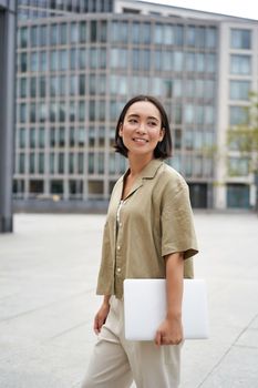 Vertical shot of asian girl walks with laptop on city street, smiles.