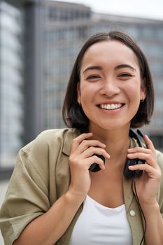 Vertical shot of beautiful asian woman posing with headphones around neck, smiling and laughing, standing on street in daylight.