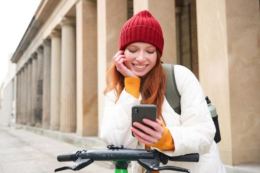 Redhead girl, tourist with backpack, uses mobile phone to rent e-scooter on streets of European city.