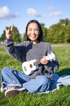 Happy asian girl playing ukulele in park, showing rock n roll, heavy metal horns sign and smiling, having fun outdoors.