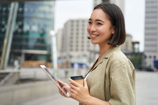 Beautiful girl standing in city square with tablet, reading and drinking takeaway coffee.