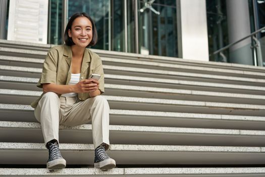 Portrait of smiling asian girl sits on stairs outdoors, sending message, using smartphone app, looking at mobile phone screen.