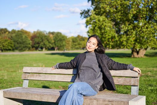 Portrait of young woman in outdoor clothes, sitting on bench relaxed, smiling and enjoying view on green park.