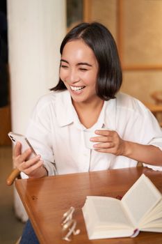 Happy asian woman sits in cafe with cup of coffee and book, answer video call on smartphone, laughing and talking via camera on mobile phone.