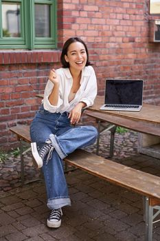 Vertical shot of good-looking asian woman with laptop, sitting in outdoor cafe, drinking coffee and smiling, laughing carefree.