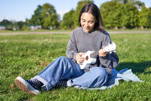 Carefree asian girl singing and playing ukulele in park, sitting on grass, musician relaxing on her free time outdoors.