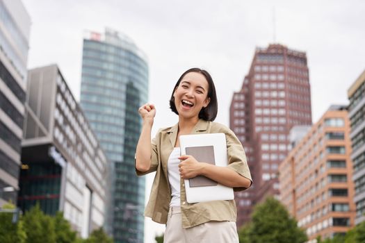 Cheerful asian girl in city centre, showing yes, hooray gesture, standing with laptop and celebrating.
