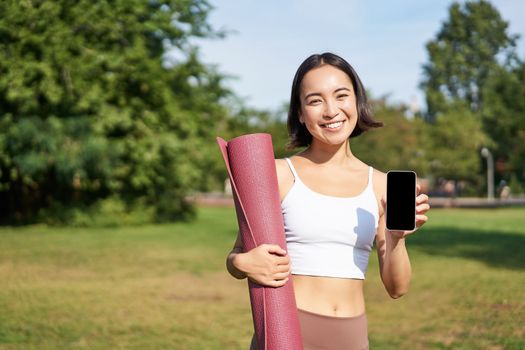 Excited fitness girl recommends application for sport and workout, shows phone screen, standing with rubber yoga mat in park after training session.