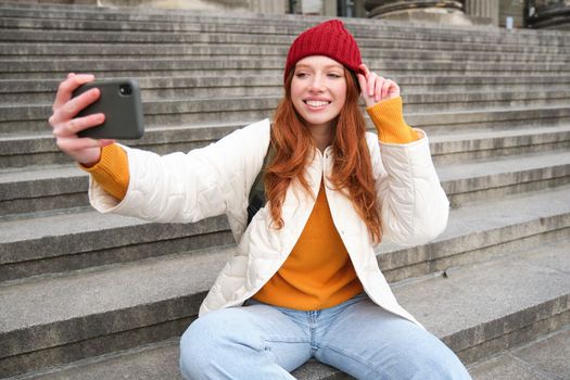 Stylish young girl in red hat, takes photos on smartphone camera, makes selfie as she sits on stairs near museum, posing for photo with app filter.