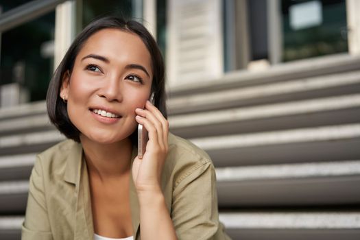 Cellular technology. Smiling asian girl talks on mobile phone, makes a call. Young woman with telephone sits on stairs.