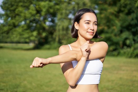 Portrait of young fitness woman stretching her arms, warm-up before training session, sport event in park, jogging and excercising.