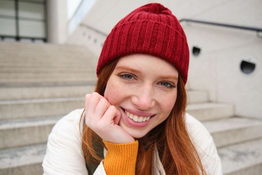 Beautiful redhead student, girl in red hat, smiles sincere, looks happy and relaxed, sits on stairs outdoors. Copy space