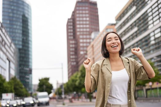 Portrait of happy asian woman, dancing and feeling joy, triumphing, raising hand up in victory gesture, celebrating on streets.