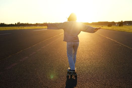 Carefree teen skater girl riding her skateboard with hands spread sideways, skating alone towards the sun.