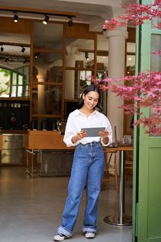 Young asian woman with tablet, standing in front of cafe entrance, inviting guests and customers, smiling broadly at camera.