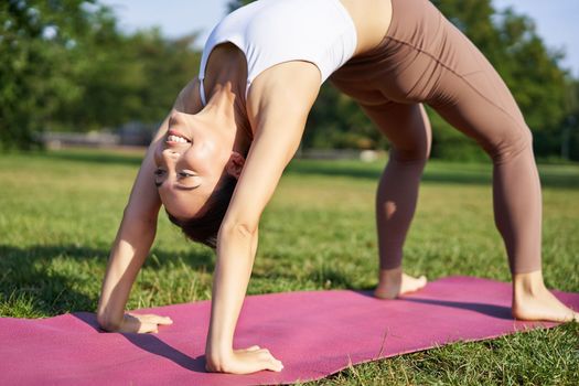Woman smiling while making bridge asana, doing yoga in park on rubber mat.