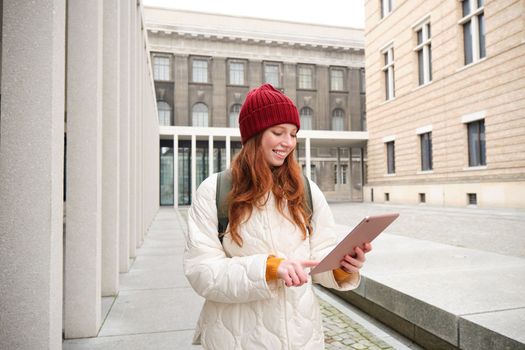 Stylish ginger girl, tourist walks with digital tablet around city, woman connects to iternet on her gadget, looking up information, texting message.