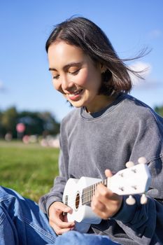 Vertical shot of smiling asian girl singing and laughing, playing ukulele, learn how to play instrument, sitting outdoors in park.